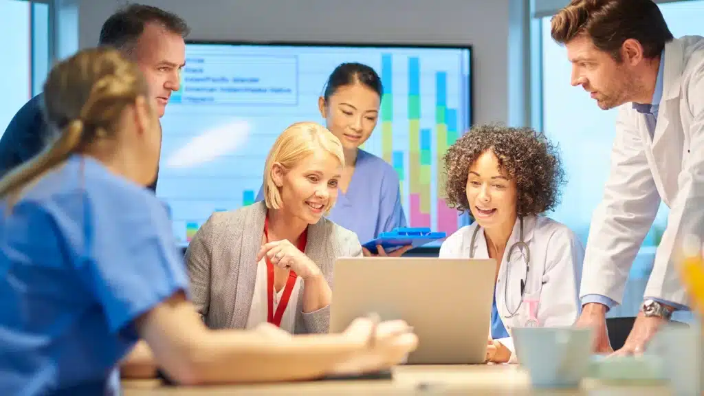 A group of doctors and professionals gathered around a table, reviewing a laptop while a bar graph is projected on the screen behind them.