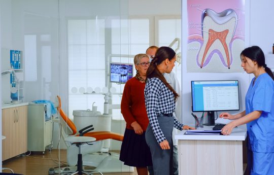 Dentist inviting senior man in consultation dental room while nurse giving to patient form to fill indicating to sit on chair in waiting area. Slow motion shot crowded professional orthodontist office